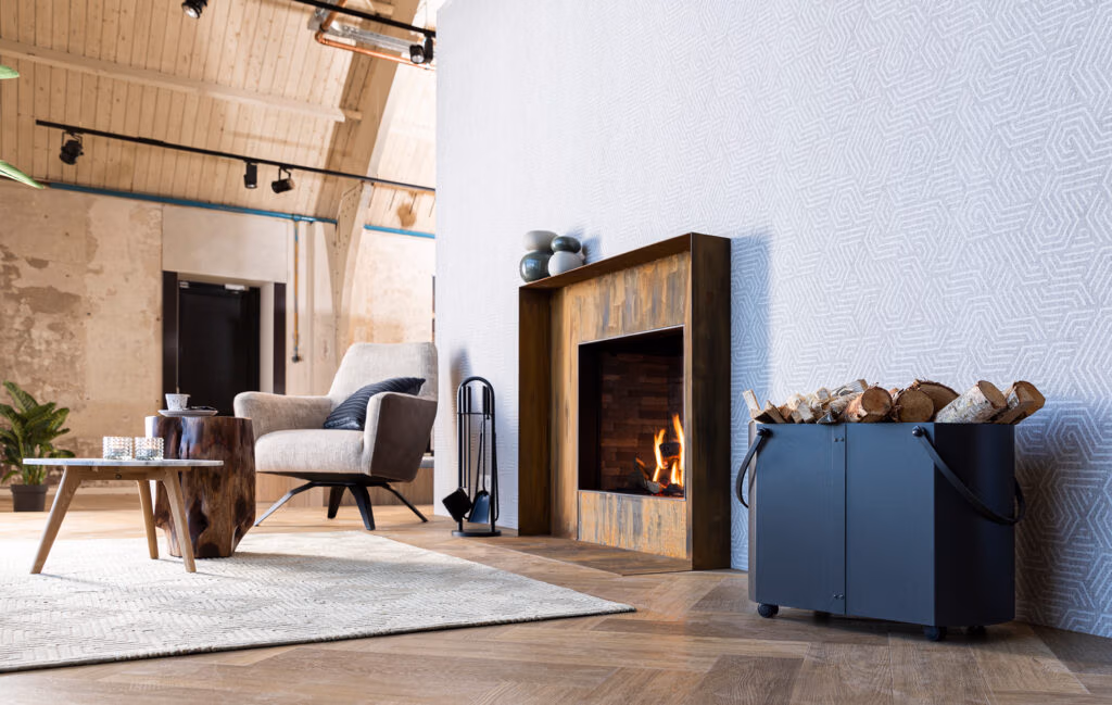Een moderne woonkamer met een verlichte open haard, grijze fauteuil, houten bijzettafel, vloerkleed met patroon en stijlvolle haardaccessoires zoals een metalen houtblokkenhouder gevuld met brandhout op een houten vloer.
