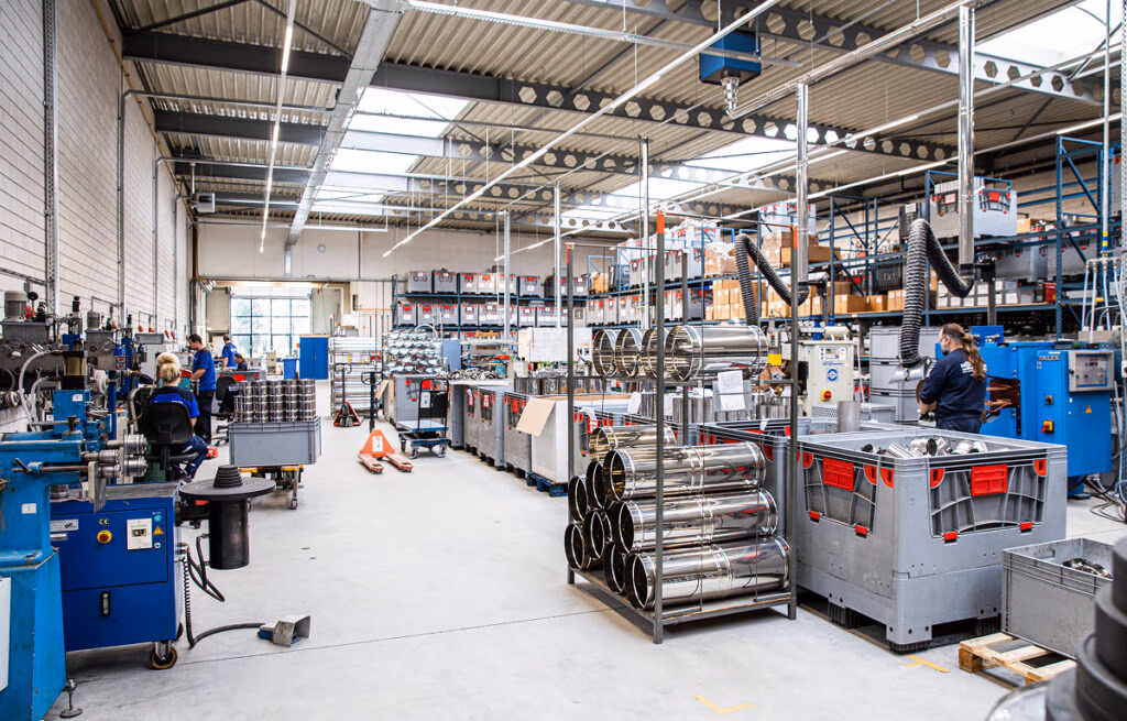 Een helder verlichte fabrieksvloer met arbeiders die machines en apparatuur bedienen, metalen cilinders, opslagbakken en planken vol met onderdelen, haardaccessoires en voorraden onder een hoog plafond met zichtbare balken en dakramen.