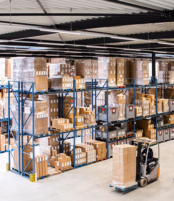 Een magazijn met hoge rekken vol dozen, haardaccessoires en pakjes. Een werknemer bedient een vorkheftruck en verplaatst gestapelde dozen op een pallet over de betonnen vloer. De ruimte is helder verlicht en georganiseerd.