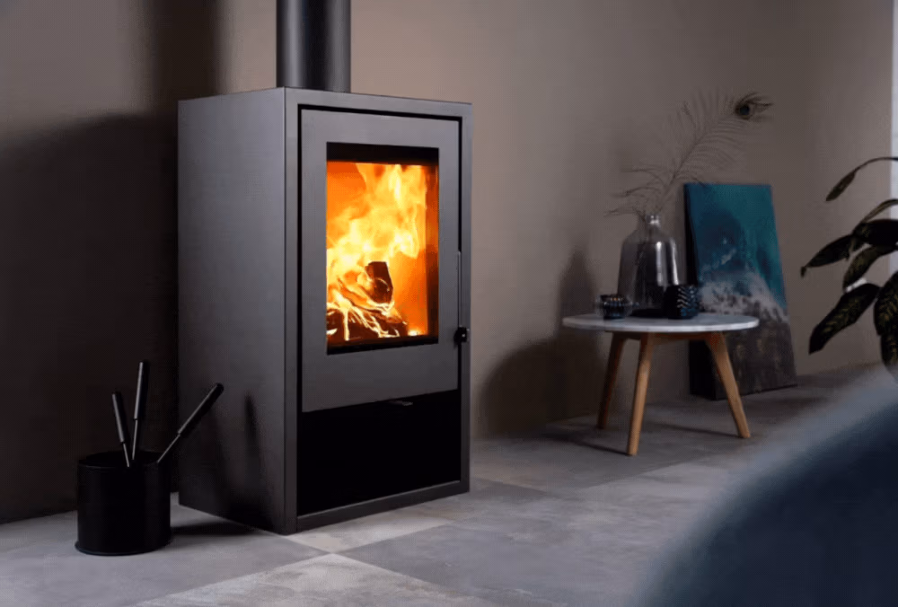 Een moderne zwarte houtkachel met een glazen deur toont een helder vuur binnenin. Haardaccessoires complementeren de minimalistische kamer, waar de kachel op een tegelvloer staat in de buurt van een kleine ronde tafel en plantendecor.