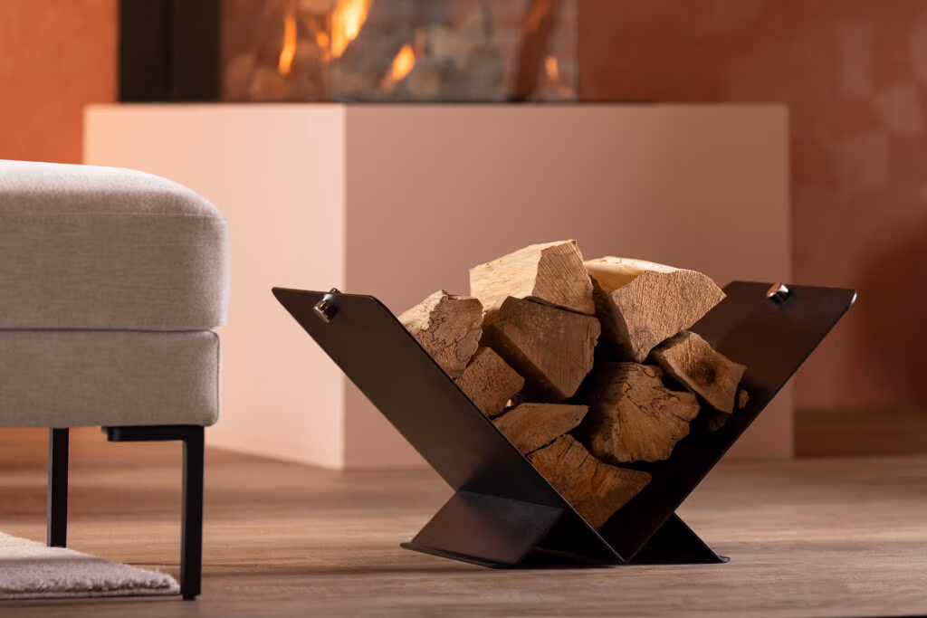 Een moderne zwart metalen haardhouthouder, een stijlvol stuk van haardaccessoires, is gevuld met gehakte houtblokken en staat op een houten vloer naast een lichtgekleurde bank, met een brandende open haard op de achtergrond.