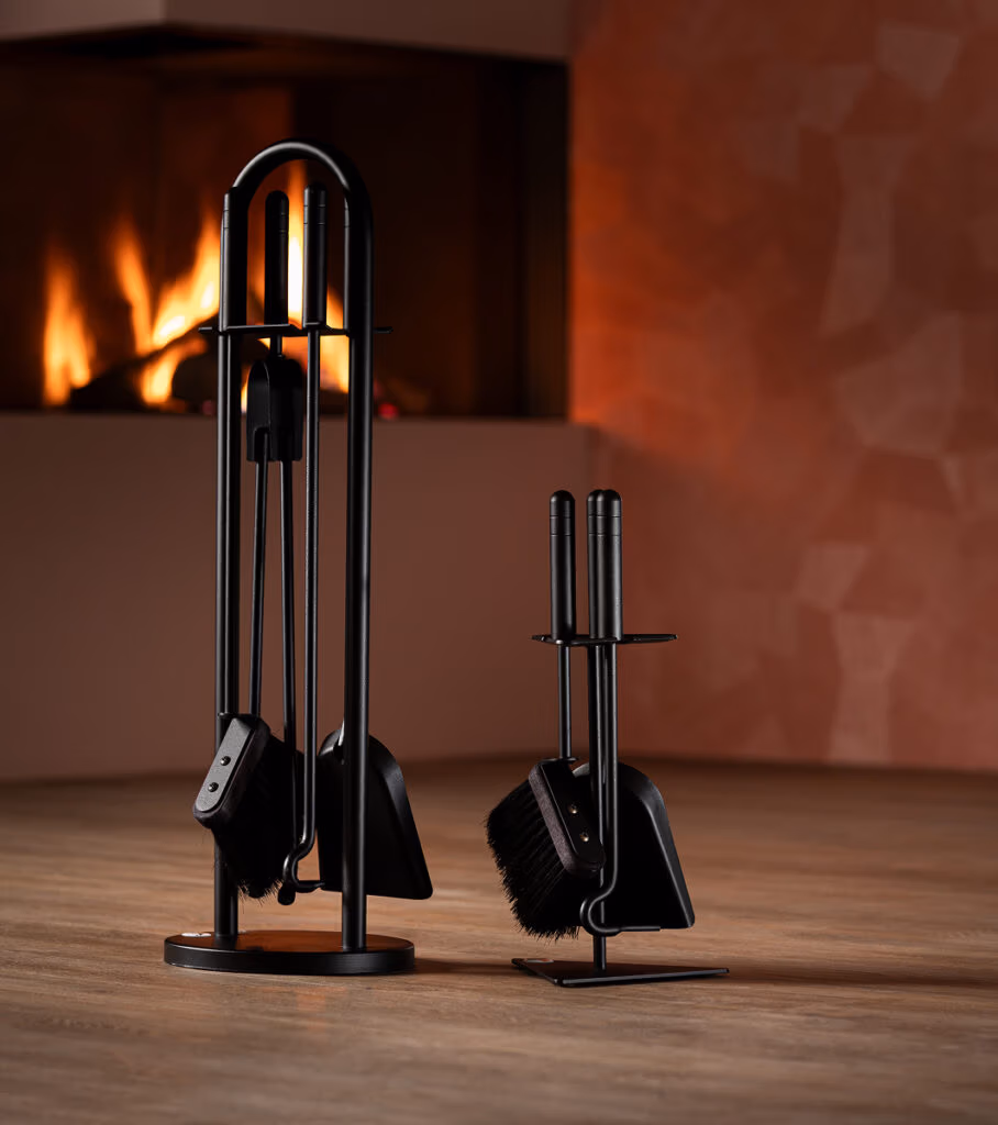 Twee zwarte haardaccessoires haardgereedschapssets rusten op een houten vloer voor een brandende open haard, met daarachter een roze muur met geometrisch patroon. De ene set is hoog en heeft vier gereedschappen, de andere is korter en heeft drie gereedschappen.