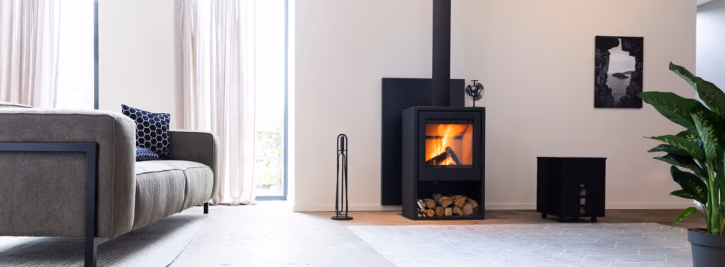 Moderne woonkamer met een brandende houtkachel en stijlvolle haardaccessoires, houtblokken eronder opgeborgen, een grijze bank met blauwe en witte kussens, een grote groene plant, transparante gordijnen en een zwart-witfoto aan de muur.