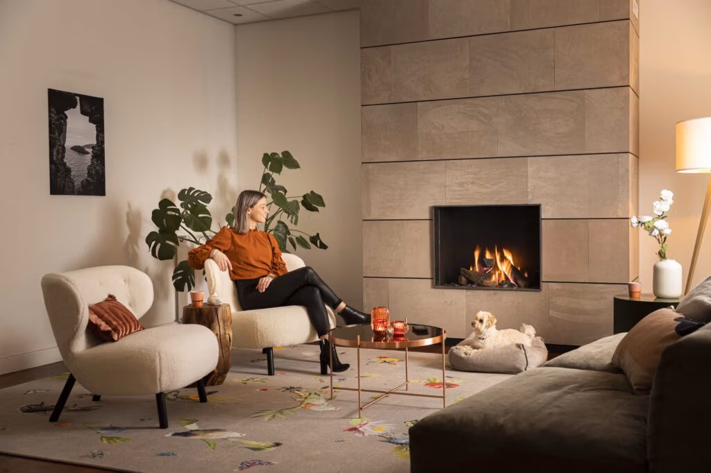 Een vrouw zit op een witte stoel bij een moderne open haard versierd met haardaccessoires in een gezellige woonkamer. Een kleine hond rust uit op een bed naast haar. De ruimte is voorzien van planten, een bank, een ronde salontafel en zachte, warme verlichting.