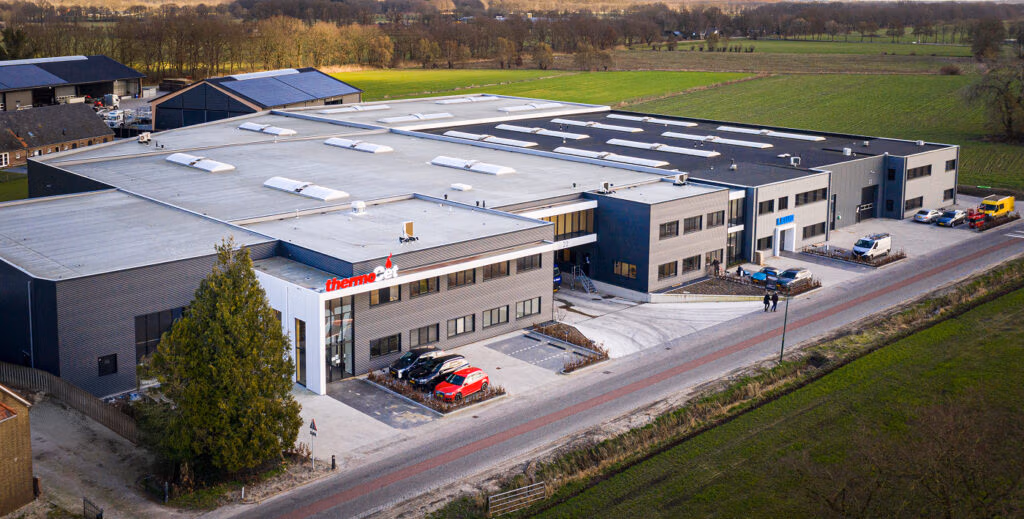 Luchtfoto van een groot industrieel gebouw met een uithangbord van ThermoNoord, omringd door auto's, groen en velden onder een gedeeltelijk bewolkte hemel - uw bestemming voor hoogwaardige haardaccessoires en meer.
