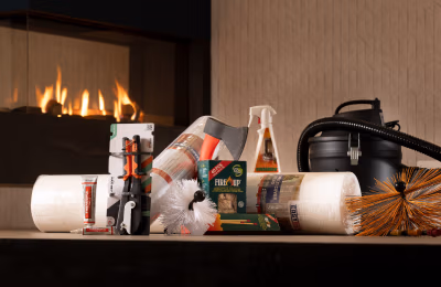 Een verscheidenheid aan producten voor het schoonmaken van open haarden en schoorstenen, waaronder borstels, sprays, houtblokken en een stofzuiger, uitgestald op een tafel met een moderne open haard die brandt op de achtergrond.
