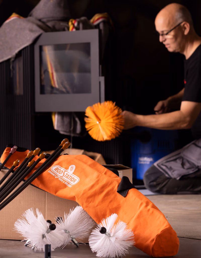 Een man knielt naast een houtkachel en houdt een oranje schoorsteenborstel vast. Op de voorgrond liggen schoonmaakborstels, staven en een oranje draagtas met de tekst ROSTFREI-essentiële Haardaccessoires om je haard schoon en veilig te houden.