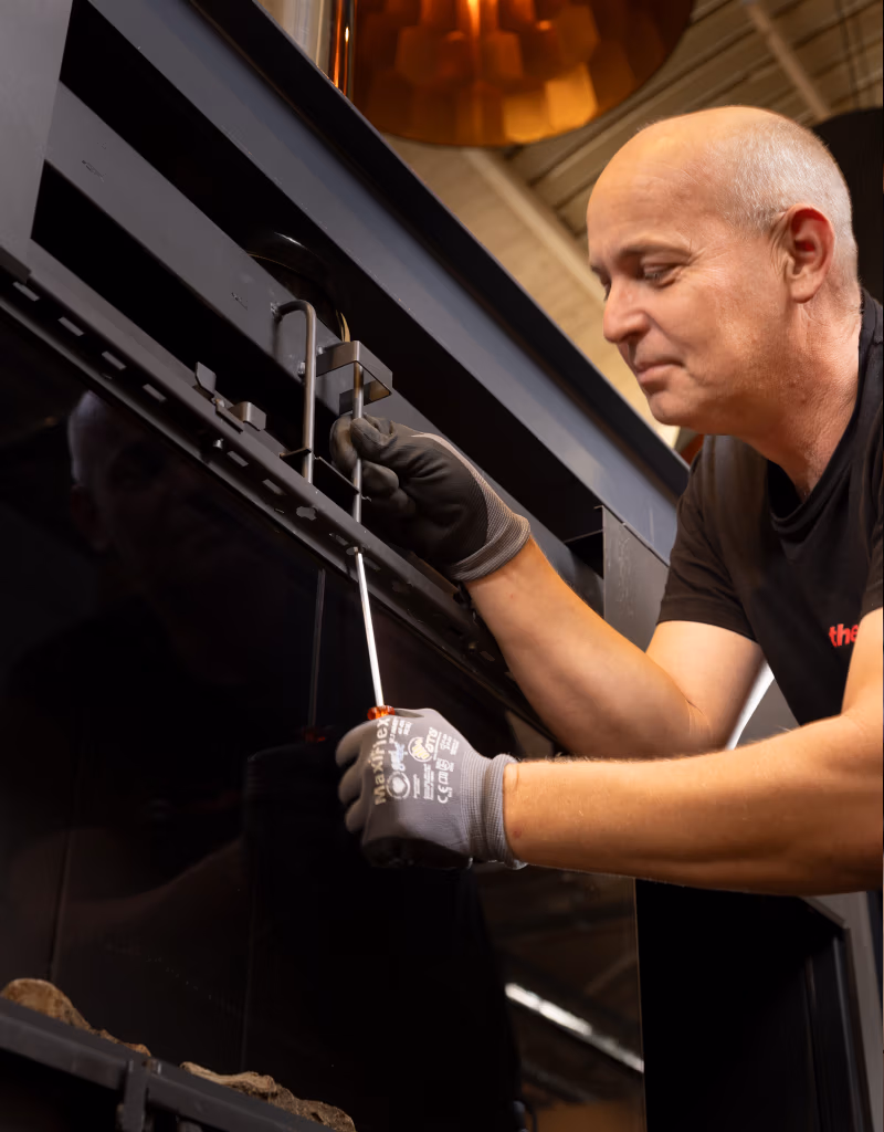 Een kale man met handschoenen aan gebruikt een schroevendraaier om een zwart metalen apparaat af te stellen, mogelijk een open haard of kachel, binnenshuis onder warme verlichting.