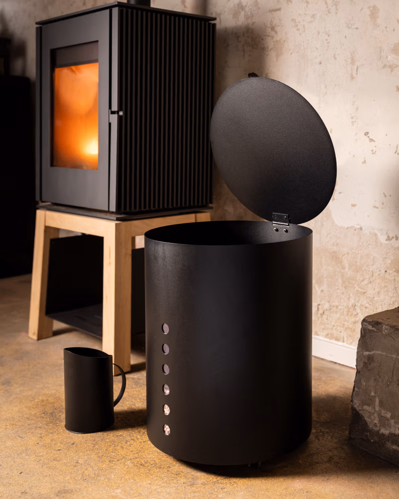 Een moderne zwarte metalen container, ideaal voor haardaccessoires, met een scharnierend deksel staat open naast een bijpassende kan op een betonnen vloer. Op de achtergrond staat een houtkachel met gloeiraam op een houten standaard tegen een muur met structuur.