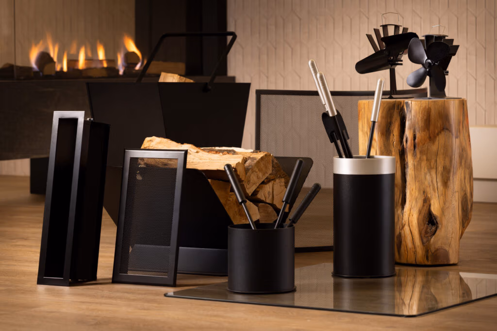 Een set moderne haardaccessoires, brandhout en gereedschap op een houten vloer bij een brandende open haard, met een houtblokhouder en een houtkachelventilator zichtbaar op de achtergrond.