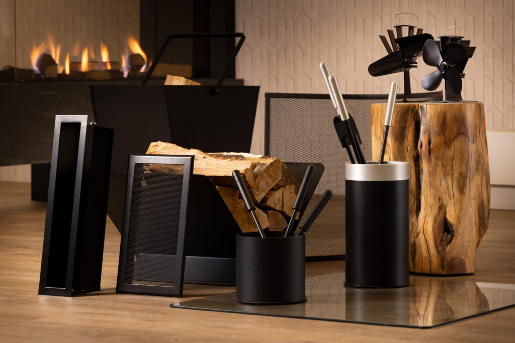 A set of modern black fireplace tools, a log holder with firewood, and a wooden block—essential haardaccessoires—are arranged on a wooden floor in front of a lit fireplace. A fan and screen are also visible in the background.