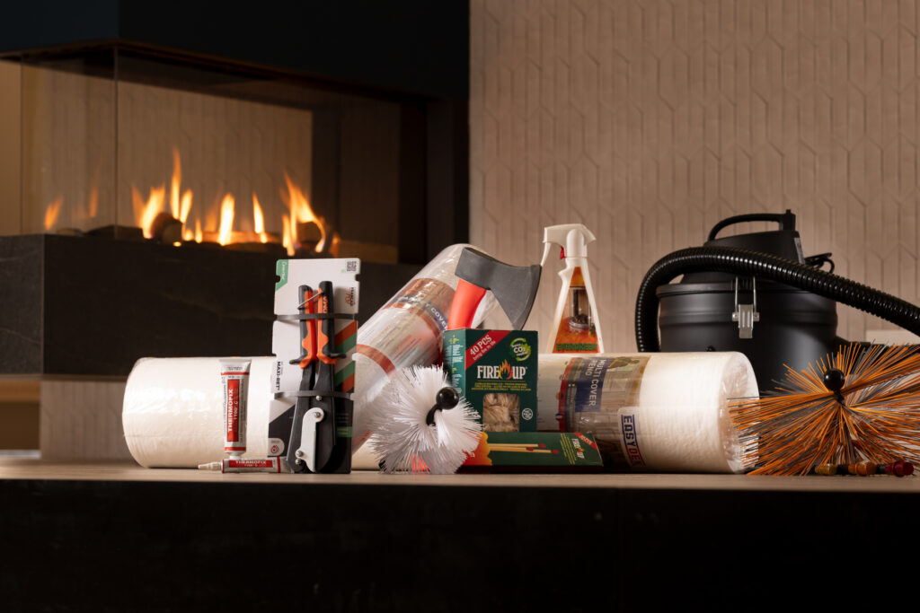A collection of fireplace cleaning supplies, including brushes, logs, cleaning spray, vacuum, and chimney sweep tools, arranged on a tabletop with a lit modern fireplace in the background.