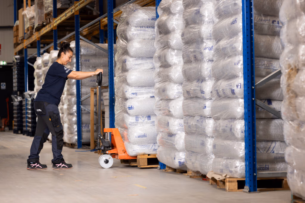 Een vrouw in werkkledij gebruikt een pallethefwagen om grote stapels verpakte goederen, waaronder haardaccessoires, te verplaatsen in een magazijn omringd door rekken met meer in plastic verpakte artikelen.