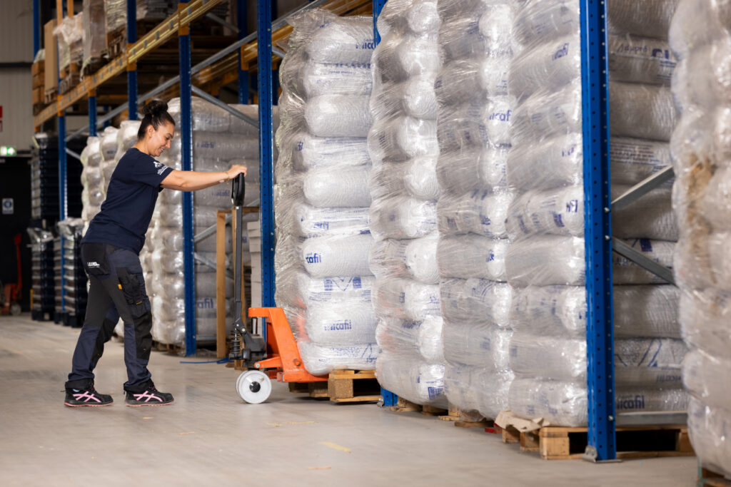 A woman in workwear uses a pallet jack to move a large stack of wrapped bags in a warehouse filled with haardaccessoires and similar stacks on shelves.