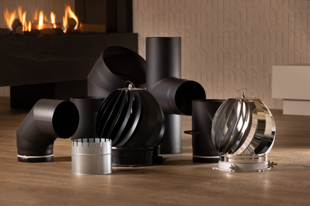 Various metal stove and chimney pipe fittings, including elbows, ducts, and vent caps, are arranged on a wooden floor indoors alongside other haardaccessoires, with a modern fireplace and textured wall in the background.