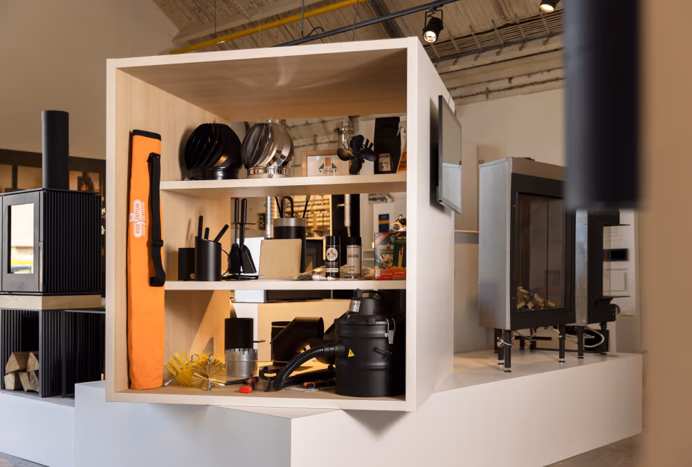 Een moderne showroom toont openhaardaccessoires en haardaccessoires op witte planken, waaronder potten, een stofzuiger en een oranje draagtas, met verschillende kachels en haarden op de achtergrond.