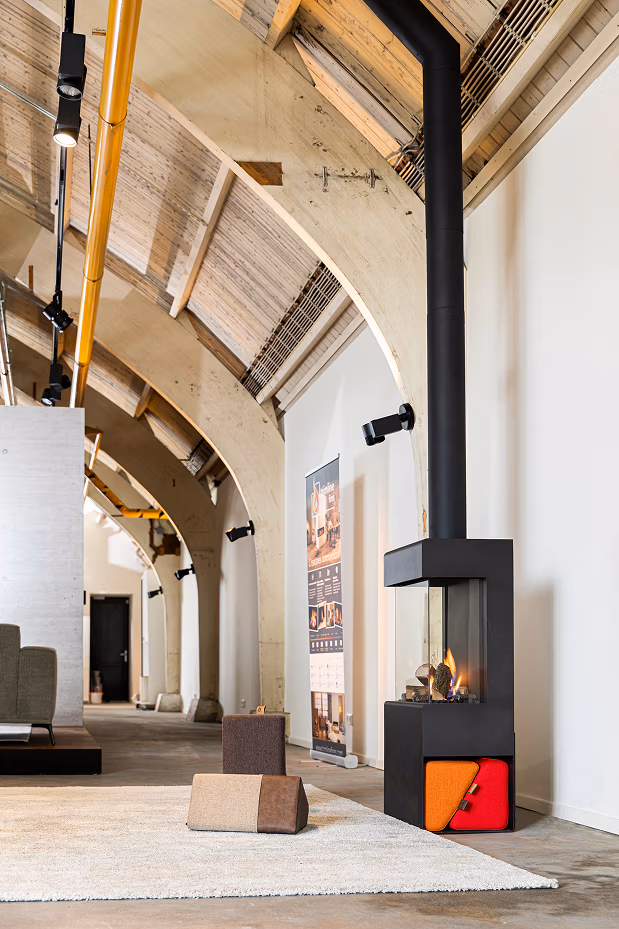 Modern interieur met een hoog, gewelfd houten plafond, een zwarte houtkachel met oranje en rode panelen, haardaccessoires netjes in de buurt, een wit vloerkleed en minimalistische bruine en bruine voetenbanken op een gepolijste betonnen vloer.