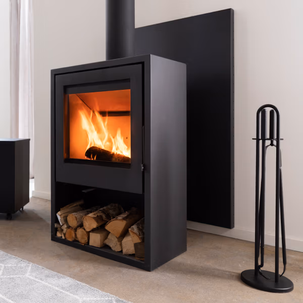 Een moderne zwarte houtkachel met een zichtbaar vuur binnenin staat tegen een lichte muur, houtblokken liggen eronder opgeslagen en stijlvolle haardaccessoires waaronder een zwart haardgereedschap staan rechts op een lichtgekleurde vloer.