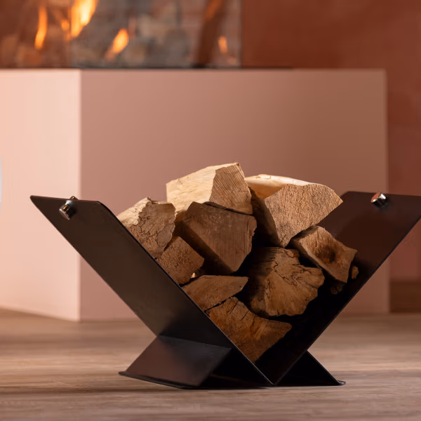Een moderne zwart metalen haardhouthouder, een stijlvol stuk van haardaccessoires, is gevuld met gehakt hout en rust op een houten vloer, terwijl een open haard warm gloeit op de onscherpe achtergrond.