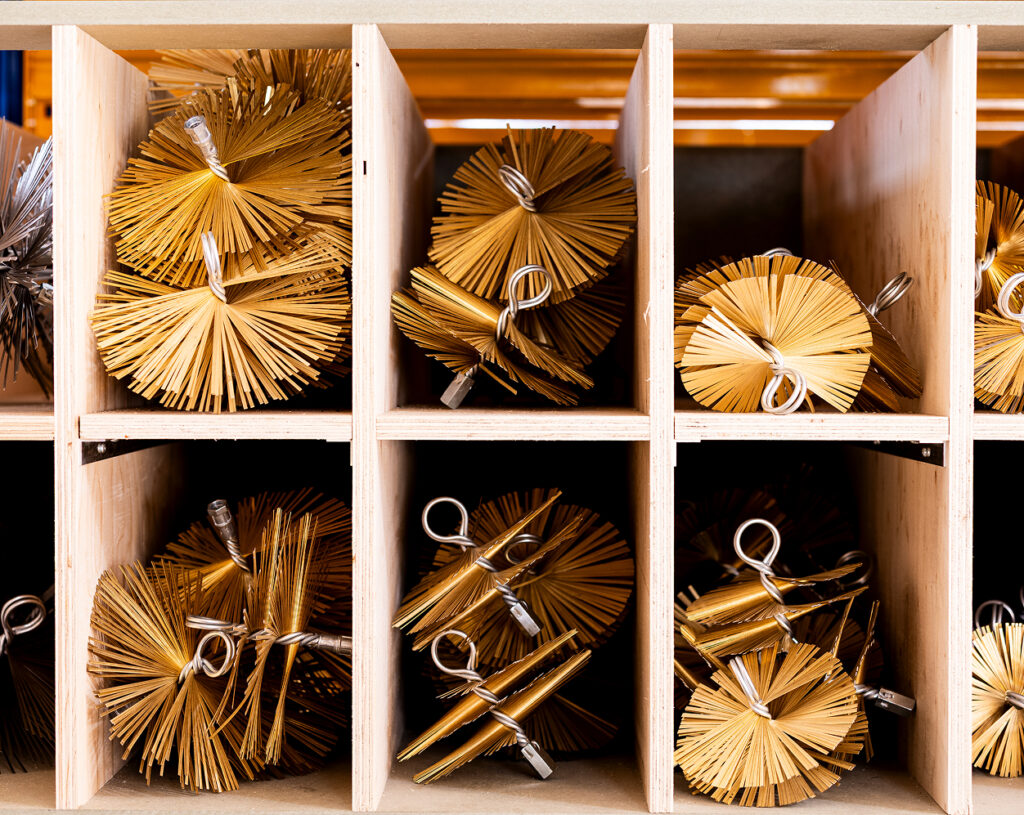 Metallic brush heads with golden bristles are neatly stored in individual wooden cubbies, each Haardaccessoires brush featuring a twisted wire handle and fanned-out bristles.