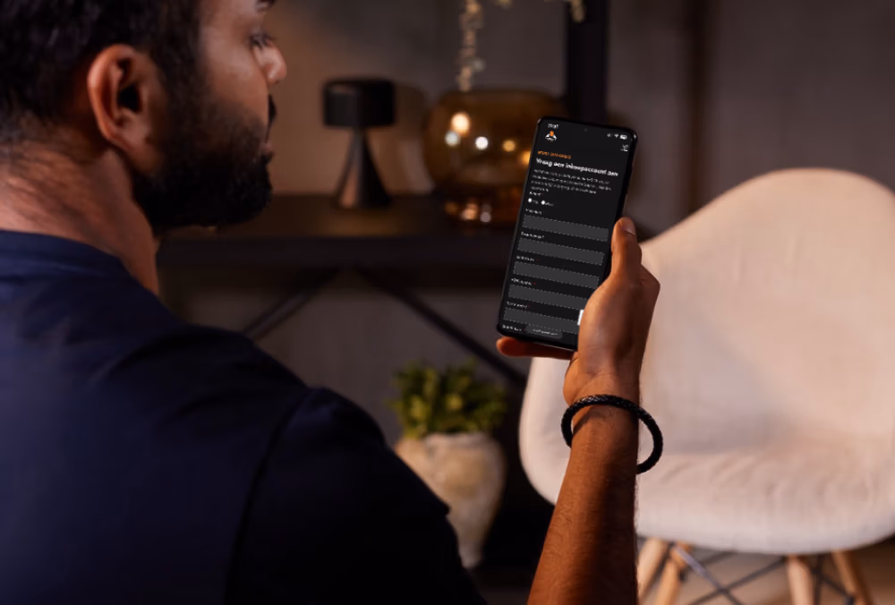 Een persoon die een smartphone vasthoudt en inhoud op het scherm leest, zittend binnen naast een witte stoel met decoratieve objecten, haardaccessoires en een potplant op de achtergrond.
