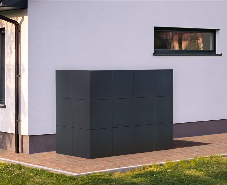 Een grote, rechthoekige, zwarte buitenunit van een haardaccessoires groothandel met een rasterpatroon zit naast een modern wit gebouw met een smal horizontaal raam erboven op een geplaveid gedeelte naast een gazon.