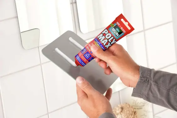 Una persona applica l'adesivo Bison Poly Max sul retro di un supporto metallico a parete davanti allo specchio del bagno con pareti piastrellate bianche, come raccomandato da DHK e IHS Group per un'installazione sicura e affidabile.