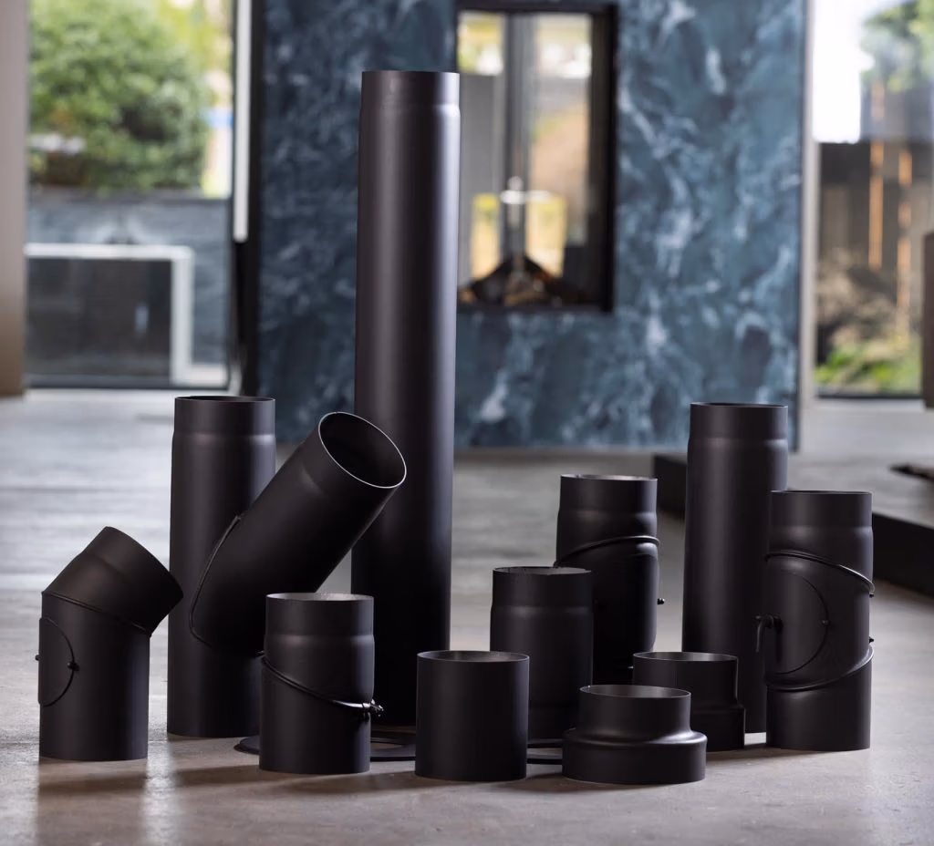 Een set zwarte metalen schoorsteenpijpen en -fittingen, afkomstig van een groothandel in haardaccessoires, staat opgesteld op een moderne betonnen vloer, met op de achtergrond een open haard en een marmeren muur.
